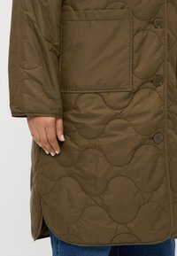 Ελαιόχρω�μο επενδυμένο παλτό με κυματιστό σχέδιο, που διαθέτει μπροστινή τσέπη, κουμπιά και στρογγυλή βάση. Μαλακή, λείες υφή.