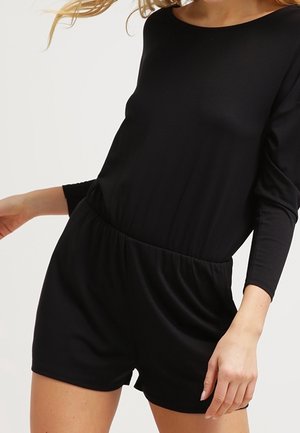 Kobieta ubrana w czarny kombinezon z długim rękawem i elastyczną talią, stojąca na tle jednolitego jasnego tła.