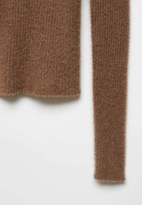 Manche et ourlet inférieur d'un pull en tricot marron pelucheux visibles sur un fond blanc, montrant un tissu à la texture douce.