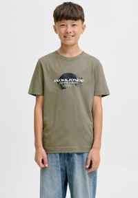 Dečko s kratkom smeškom smešnom kose noseći maslinasto zelenu majicu Jack & Jones i plave traperice, stoji ispred jednostavne bijele pozadine.