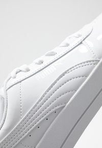 Baskets en cuir blanc verni présentant des panneaux latéraux texturés, un bout lisse et des lacets plats blancs. Logo affiché sur le côté.