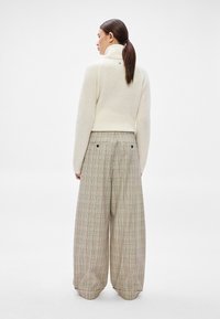 Camisola de malha creme com gola alta, combinada com calças largas de padrão xadrez em bege, castanho e detalhes subtis em vermelho. Vista de costas, design minimalista.