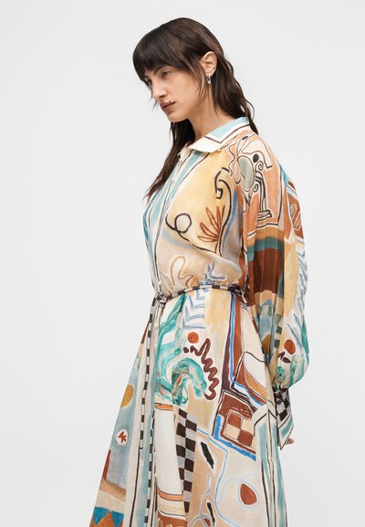 Γυναίκα με μακριά σκούρα μαλλιά που φοράει πολύχρωμο φόρεμα με αφηρημένο μοτίβο, φαρδιά μανίκια και λεπτή ζώνη, κοιτάζοντας προς τα αριστερά σε απλό φόντο.