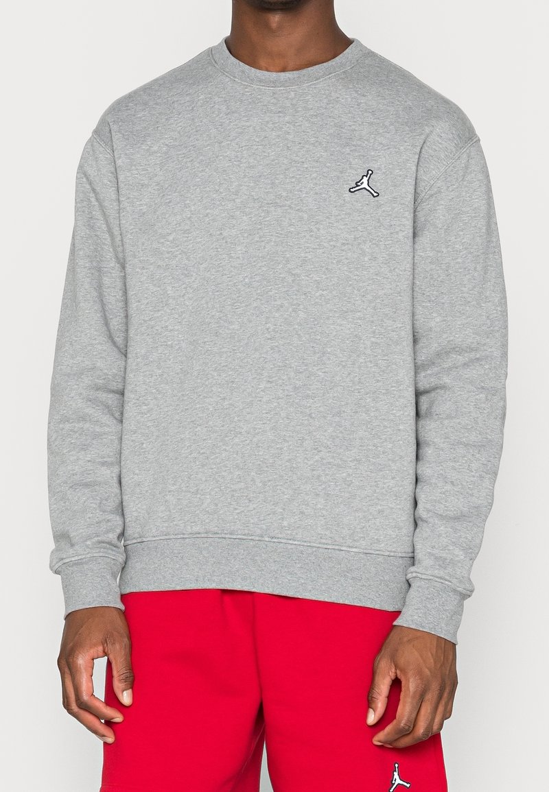 Homme portant un sweat-shirt gris clair à col rond et un short rouge, tous deux avec un petit logo Jumpman noir et blanc.