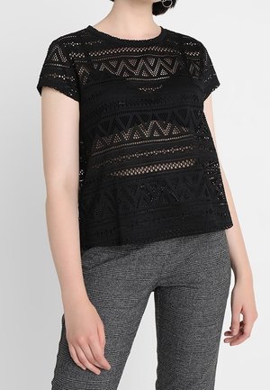 Czarny top z krótkim rękawem z wzorzystymi wycięciami, z okrągłym dekoltem, zestawiony z szarymi spodniami w kratę o wysokim stanie.