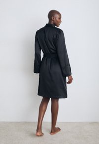 Svart satinrock med långa ärmar och bälte, med krage, slät yta och knä-längd. Baksidan är enkel utan utsmyckningar.