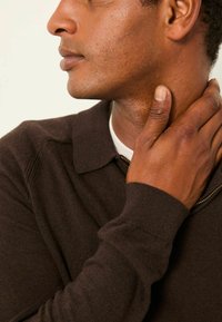 Braunes Strickpolo mit Kragen, ausgestattet mit einem Reißverschluss und gerippten Bündchen, dargestellt mit einer Hand, die den Halsbereich berührt.