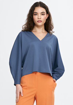 Niebieska bluzka w serek z długimi, luźnymi rękawami, wykonana z gładkiego materiału, w połączeniu z pomarańczowymi spodniami. Posiada prosty dół i szwy boczne.