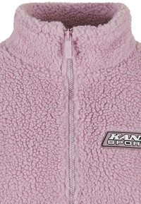 Chaqueta de forro polar rosa con cremallera y cuello alto, que presenta una tela texturizada y un logotipo negro "KANDI SPORT" en la esquina inferior derecha.