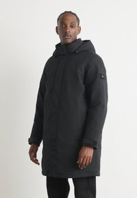 Schwarze Kapuzenparka mit Reißverschluss, weichem Material und verstellbaren Bündchen. Mit einem Logoschild auf dem linken Ärmel.