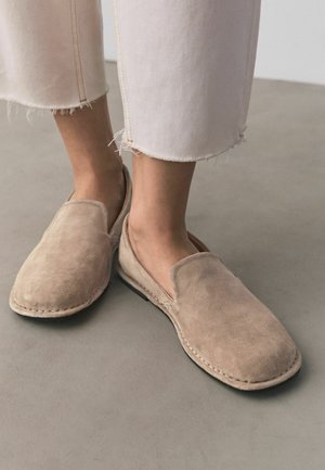 Feet wearing beige suede slip-on shoes with visible stitching, paired with cream-colored cropped frayed-edge pants on a gray floor.
