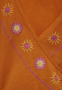 Orange tyg med broderade motiv av sol och blommor i gult, rosa och lila, med dekorativ sömnad längs sömmarna.