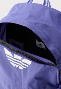 Μωβ υφασμάτινο σακίδιο με μεγάλη άνοιγμα, με λευκό λογότυπο στο μπροστινό μέρος και μαύρη επένδυση εσωτερικά. Διακρίνονται οι ραφές.