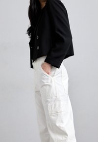 Blazer negro corto con dos botones y bolsillos laterales, combinado con pantalones cargo blancos que presentan costuras visibles y un detalle de bolsillo lateral.