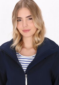 Giacca blu navy con zip e cappuccio foderato in pile, abbinata a una maglietta bianca a righe. Materiale morbido e design snellito con cerniere.