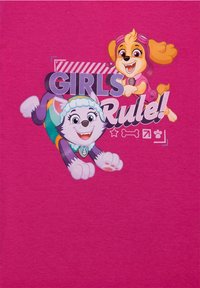 Maglietta rosa con cani dei cartoni, uno con un mantello giallo e occhiali rosa, e l'altro con un mantello bianco e una sciarpa blu. Il testo dice "Le RAGAZZE comandano!"