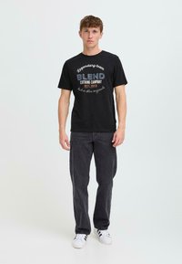 Juodas medvilnės marškinėliai su "BLEND Clothing Company" grafika ir įkurtuves data, atspausdinta balta ir mėlyna spalvomis. Dėvima su tamsiai pilkomis kelnėmis ir baltomis sportinėmis bateliais.