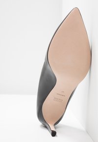 Chaussure à talon haut en cuir noir à bout pointu reposant sur une surface blanche, semelle visible avec les marques de la marque et de la taille.