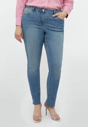 Mujer con vaqueros pitillo azul claro, camisa de rayas rosa y blanca, y tacones blancos con detalles de cuentas plateadas.