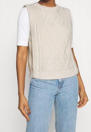 Henkilö pukeutuneena kermanväriseen palmikkoneuletakkeeseen, jonka alla on valkoinen lyhythihainen paita ja vaaleansiniset korkeavyötäröiset farkut.