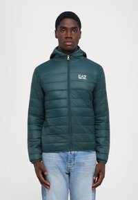 Grøn polstret jakke med lynlås foran, hætte og EA7-logo. Quiltet design med glat tekstur og tætsiddende silhuet. Jeans synlige nedenfor.