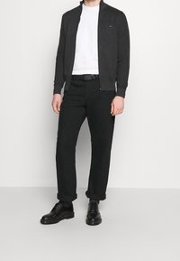 Felpa grigia scura con zip sopra una maglietta bianca, abbinata a pantaloni neri con risvolto e scarpe nere stringate. Design semplice, senza motivi.