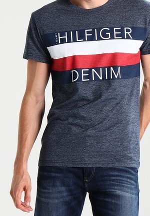 Mężczyzna w granatowej koszulce Tommy Hilfiger Denim z białymi, czerwonymi i niebieskimi poziomymi paskami oraz ciemnoniebieskich dżinsach.