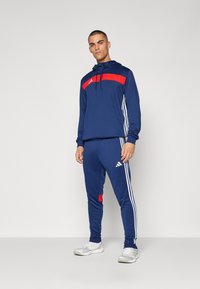 Survêtement bleu marine avec des accents rouges, comprenant un haut à capuche et un pantalon fuselé. Rayures blanches sur les côtés et détail du logo sur la poitrine.