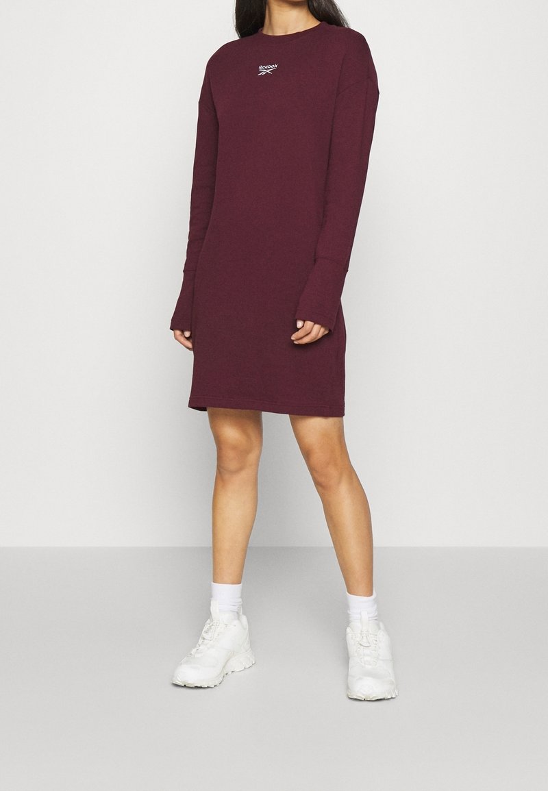 Femme portant une robe Reebok bordeaux à manches longues et longueur au genou, avec des chaussettes blanches et des baskets blanches, debout devant un fond uni.