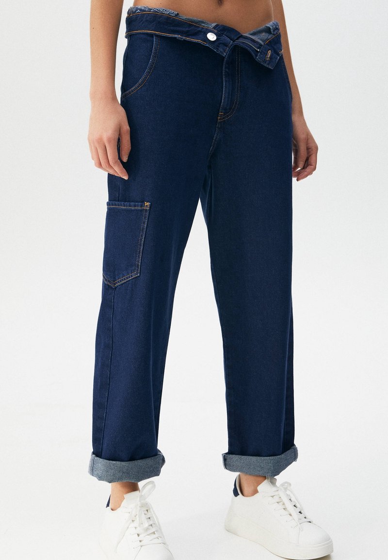 Personne portant un jean bleu foncé retroussé avec une poche latérale et des baskets blanches, debout devant un fond blanc uni.