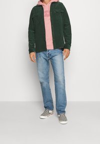 Chemise en velours côtelé vert portée sur un sweat à capuche rose, associée à un jean bleu et des baskets à carreaux noir et blanc. Tenue décontractée et superposée.
