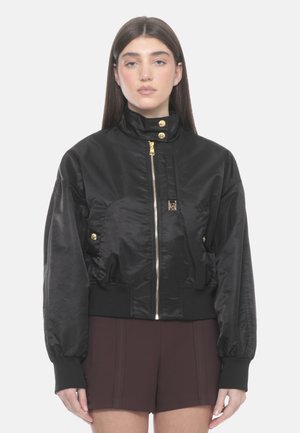 Giovane donna che indossa una giacca bomber nera con zip e bottoni dorati e pantaloncini marrone scuro, in piedi davanti a uno sfondo chiaro e uniforme.