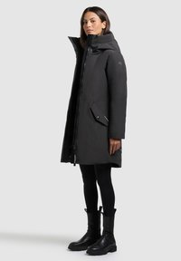 Svart vadderad jacka med hög krage, dragkedjestängning, sidofickor och en slät, smooth yta. Bärs över svarta leggings och kraftiga svarta stövlar.