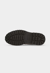 Semelle de chaussure en caoutchouc noir avec des rainures profondes et larges pour une meilleure adhérence et durabilité, présentée sur un fond gris clair uni.