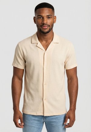 Hombre con barba corta que lleva una camisa beige de manga corta con botones y unos vaqueros azul claro, de pie contra un fondo claro y sencillo.