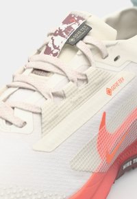 Chaussure de course légère avec une tige en mesh blanc, des accents roses, un logo Gore-Tex et des superpositions texturées. Dispose d'une semelle rose avec une bonne adhérence.