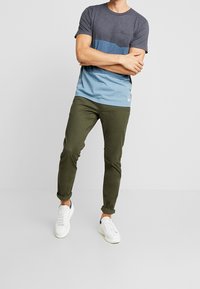 Tričko s krátkým rukávem v námořnické a světle modré barvě, s náprsní kapsou; kombinováno s olivově zelenými chinos kalhotami a bílými teniskami.