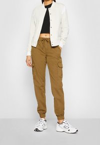 Jasnoszara kurtka bombowca z białymi akcentami na ramionach, połączona z beżowymi spodniami cargo z bocznymi kieszeniami. Całość dopełniają białe sneakersy.