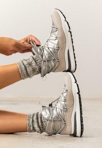 Zapatillas altas plateadas y beige con un acabado texturizado, cordones elásticos y una suela blanca. Llevadas con calcetines gris de punto grueso.