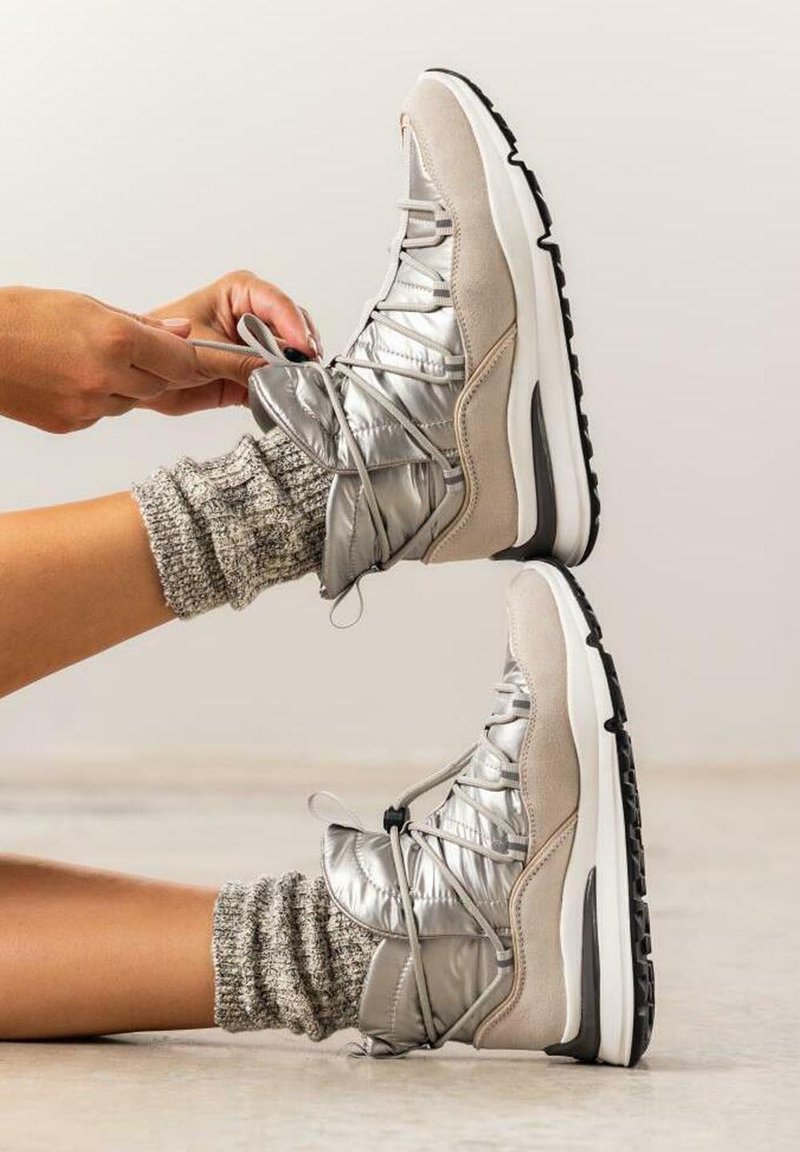Zapatillas altas plateadas y beige con un acabado texturizado, cordones elásticos y una suela blanca. Llevadas con calcetines gris de punto grueso.