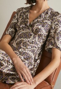 Vrouw zittend in een bruine stoel, gekleed in een kort mouw bloemenjurk met witte, gele en paarse bloemen en een gerimpelde V-hals kraag.