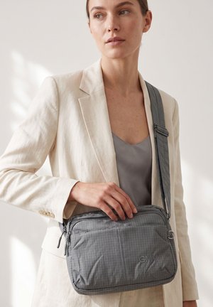 Femme en blazer beige et haut gris tenant un sac bandoulière gris avec une sangle réglable et des compartiments zippés.
