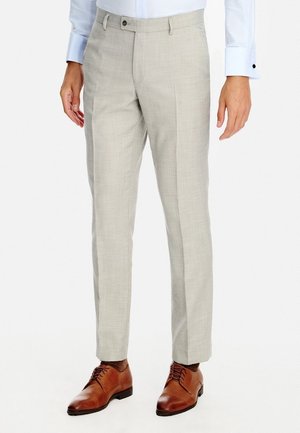 Mężczyzna noszący jasnobeżowe eleganckie spodnie i brązowe skórzane buty wizytowe, w białej koszuli z długim rękawem, stojący na białym tle.