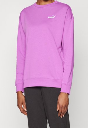 Personne portant un sweat-shirt Puma violet clair à manches longues et un pantalon noir, debout devant un fond clair uni.