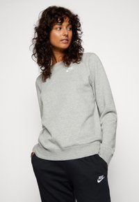 Sweatshirt Nike en tissu gris clair, avec un col rond, des poignets côtelés et un logo blanc sur la poitrine. Associé à un pantalon noir.