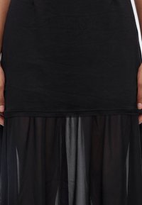 Gros plan d'une personne portant une robe noire avec un haut opaque et une jupe en tissu transparent, les mains reposant sur les côtés.