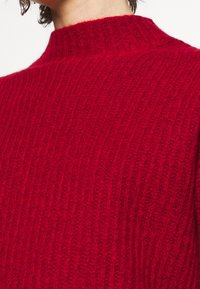 Punainen neulottu korkean kauluksen villapaita, jossa on teksturoitu resorikuviointi. Lähikuva korostaa kankaan lämpöä ja yksityiskohtia.