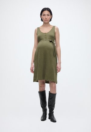 Έγκυος γυναίκα που φοράει αμάνικο ελιάς φόρεμα με κουμπιά και δέσιμο στη μέση, συνδυασμένο με μαύρες μπότες μέχρι το γόνατο, στέκεται μπροστά σε λευκό φόντο.