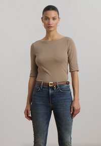 Top bege de manga longa com decote largo combinado com jeans azuis de cintura alta. Cinto castanho com fivela de latão como destaque. Textura de tecido suave.