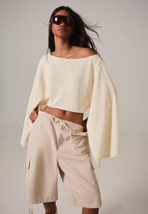 Femme portant de grandes lunettes de soleil foncées, un pull court ample crème avec de larges manches et un short beige à cordon sur fond uni.